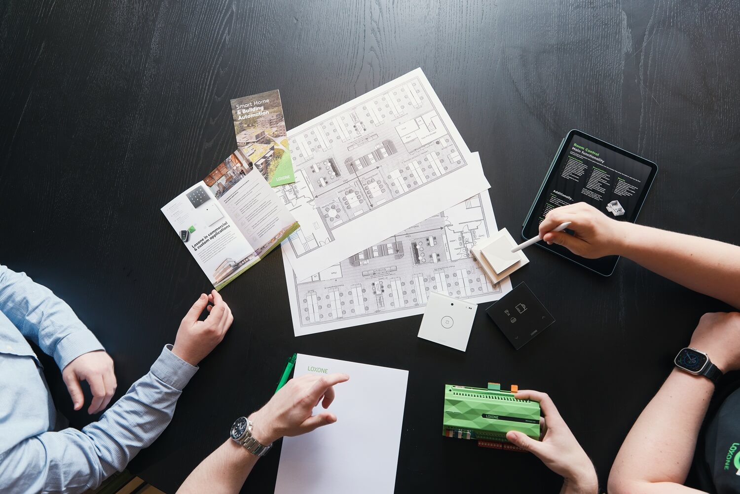 Two men and a woman discussing plans for a residential building automation system with the Loxone Touch Pure and a floor plan on the table 